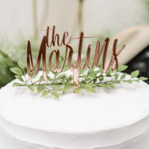 May include: A white wedding cake with a rose gold cake topper that says "the Martins" and greenery.