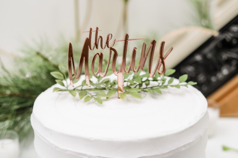 May include: A white wedding cake with a rose gold cake topper that says "the Martins" and greenery.