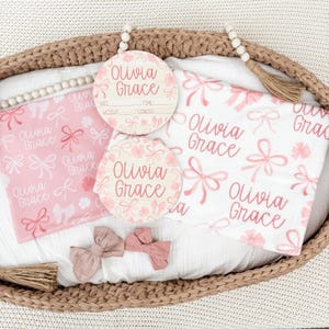 May include: A baby gift set featuring a pink and white blanket and two wooden announcement discs, all with the name "Olivia Grace." The blanket has a bow pattern. Two pink bows are also included.