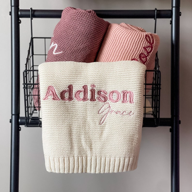 K&ouml;nnte beinhalten: Eine wei&szlig;e gestrickte Babydecke mit dem Namen "Addison Grace", der in rosa gestickt ist. Die Decke h&auml;ngt an einem schwarzen Metallgestell.