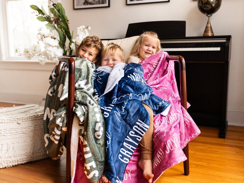 May include: Three children are sitting in a chair, each wrapped in a personalized blanket. The blankets are green, blue, and pink, with the names "MATTHEW", "GRAYSON", and "INDIVIDUALLY JOY" written in white script.