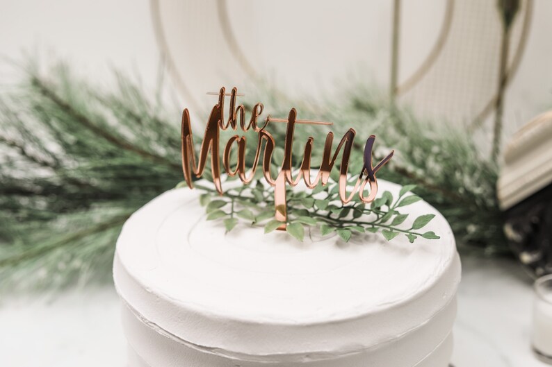 May include: A white cake with a rose gold cake topper that says "the Martins".