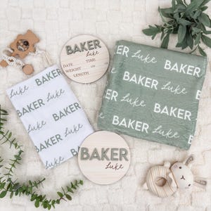 May include: A collection of baby items, including a white and a sage green blanket, both with the name "Baker" and "Luke" printed on them. There are also two wooden round signs with the name "Baker" and "Luke" on them, and a wooden baby rattle.