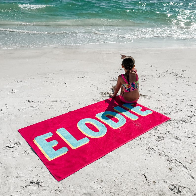 May include: A pink beach towel with the name "ELODIE" printed in large, blue letters with a yellow outline. A young person is sitting on the towel, facing away from the camera, on a white sandy beach.