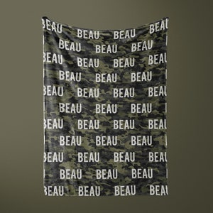 K&ouml;nnte beinhalten: Eine gr&uuml;n-schwarze Tarnmuster-Decke mit dem Namen "BEAU", der in wei&szlig;er Schrift wiederholt wird.