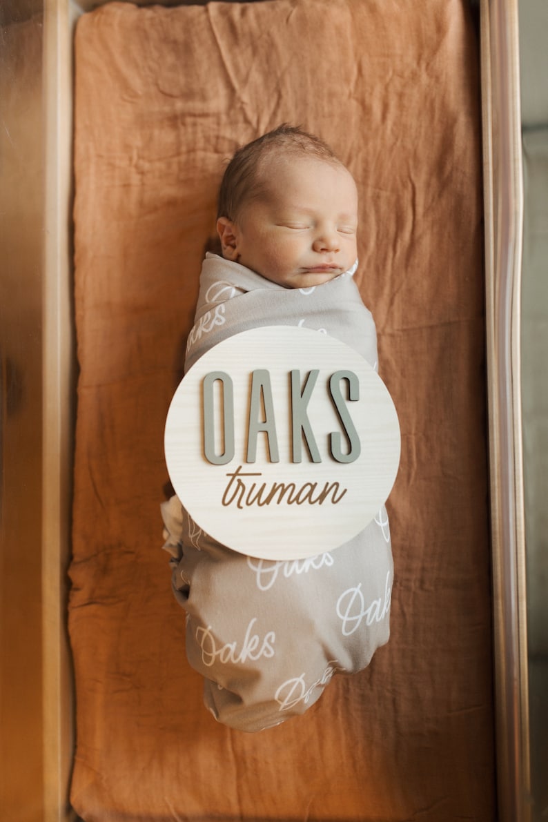 May include: A newborn baby is swaddled in a gray blanket with white lettering that says "Oaks". A round wooden sign with the name "Oaks Truman" is placed on the blanket. The baby is sleeping in a wooden bassinet with a brown fabric lining.
