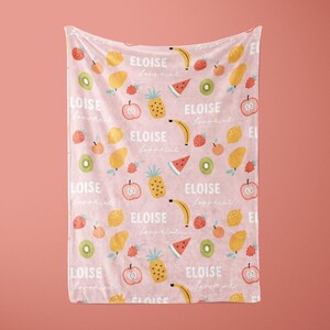 May include: Pink baby blanket with a repeating pattern of fruit, including pineapples, lemons, bananas, watermelons, kiwi, and apples. The text "ELOISE loves fruit" is repeated throughout the blanket.