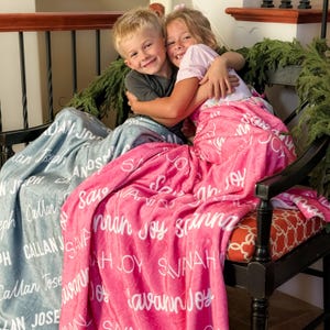 May include: Two children are embracing, seated on a black chair, each wrapped in a personalized blanket. One blanket is light blue, the other is pink, both with white script names. The chair has an orange patterned cushion and is decorated with greenery.