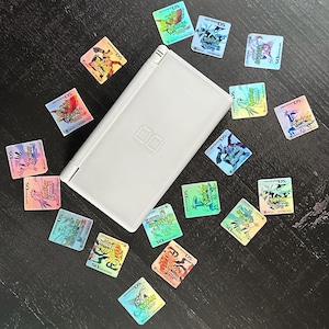Puede incluir: Una consola de juegos portátil Nintendo DS blanca rodeada de cartuchos de juego coloridos. Los cartuchos presentan varios personajes de Pokémon y el texto "DS". El fondo es una superficie oscura.