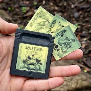 以下が含まれることがあります： 黒いゲームボーイカラーのカートリッジに、黄色いラベルが貼ってあり、「ポケットモンスター」と日本語で書かれています。カートリッジは手に持たれており、同じ日本語のテキストとポケモンのキャラクターの画像が描かれた黄色のトレーディングカードが3枚あります。