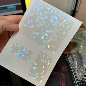 May include: A white rectangular sheet featuring four distinct adhesive stickers with a holographic shattered glass pattern. The stickers display reflective shards of light in various colours, including blue, green, and orange, against a translucent base.