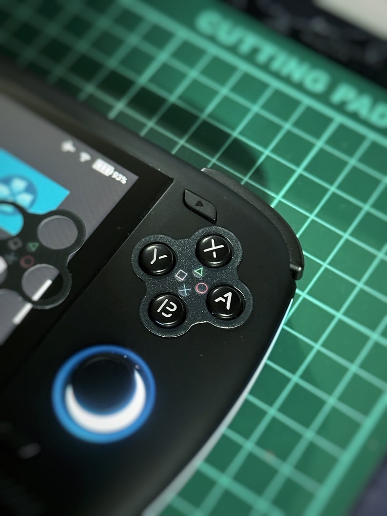 Op de afbeelding: Een zwart draagbaar gameapparaat met een blauwe en witte ronde knop en een directionele pad met vier knoppen. Het apparaat heeft een scherm dat "100%" weergeeft.