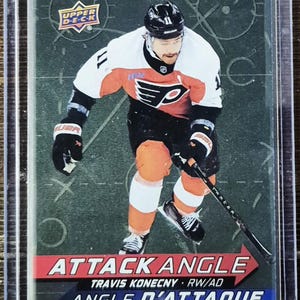 May include: A hockey trading card featuring Travis Konecny in a Philadelphia Flyers uniform. The card has the text "ATTACK ANGLE" and "ANGLE D'ATTAQUE". The player is wearing a black helmet, white jersey with orange accents, and black gloves.