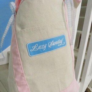 Puede incluir: Un delantal beige con paneles laterales verticales rosas y lazos de cinta de cuadros vichy azul claro y blanco. Un parche rectangular azul está centrado en el bolsillo delantero con el texto cursivo blanco Lazy Sunday impreso en el centro.