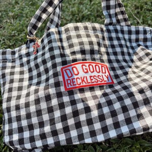 Puede incluir: Un bolso de tela con estampado de cuadros vichy en blanco y negro, con dos asas superiores y un parche rectangular de borde rojo y fondo azul claro. El parche muestra el texto DO GOOD RECKLESSLY en letras rojas. Un pequeño dije de cruz roja cuelga.