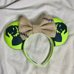 May include: Green mouse ears with a black silhouette of Jack Skellington from The Nightmare Before Christmas. The ears are decorated with a brown burlap bow.