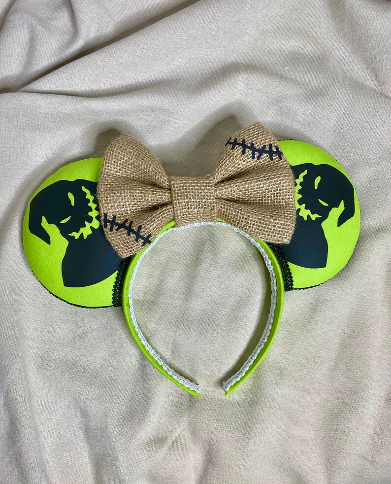 May include: Green mouse ears with a black silhouette of Jack Skellington from The Nightmare Before Christmas. The ears are decorated with a brown burlap bow.