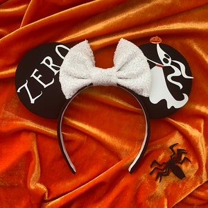 May include: Black and white Halloween-themed mouse ears with a white sequin bow. One ear features the word "ZERO" and the other has a ghost character. A black spider decoration is also present.