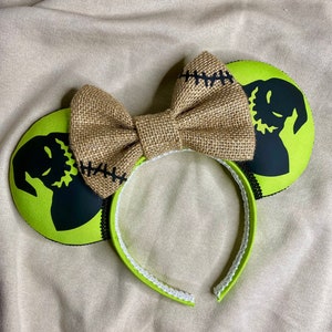 May include: Green and black mouse ears with a burlap bow. The ears feature a silhouette of the character Jack Skellington from the movie The Nightmare Before Christmas.