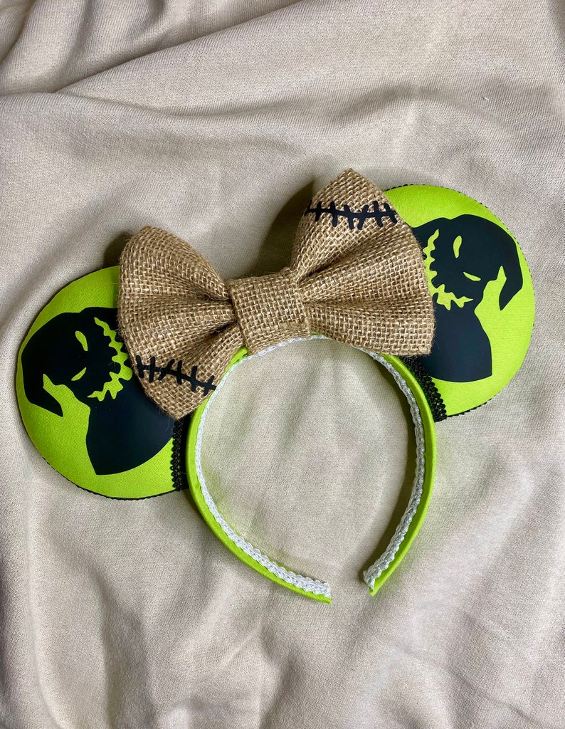 May include: Green and black mouse ears with a burlap bow. The ears feature a silhouette of the character Jack Skellington from the movie The Nightmare Before Christmas.