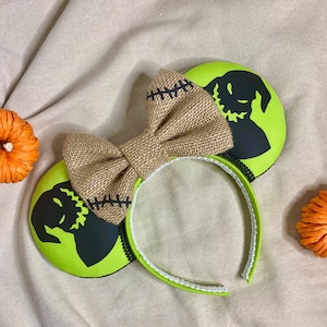 May include: Green and black mouse ears with a burlap bow. The ears feature a silhouette of the Nightmare Before Christmas character Jack Skellington.