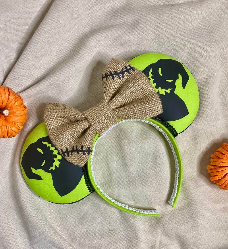 May include: Green and black mouse ears with a burlap bow. The ears feature a silhouette of the Nightmare Before Christmas character Jack Skellington.