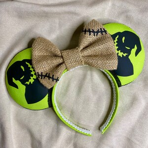 May include: Green and black mouse ears with a burlap bow. The ears feature a silhouette of the character Jack Skellington from the movie The Nightmare Before Christmas.