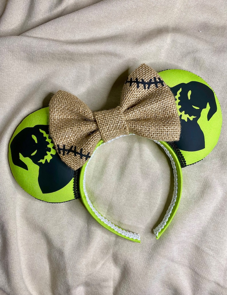 May include: Green and black mouse ears with a burlap bow. The ears feature a silhouette of the character Jack Skellington from the movie The Nightmare Before Christmas.