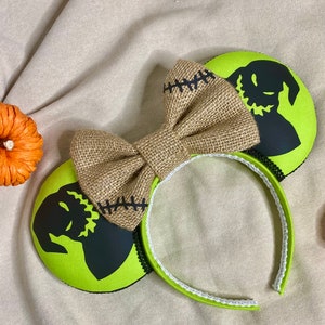 May include: Green and black mouse ears with a burlap bow. The ears feature a silhouette of the character Jack Skellington from the movie The Nightmare Before Christmas.