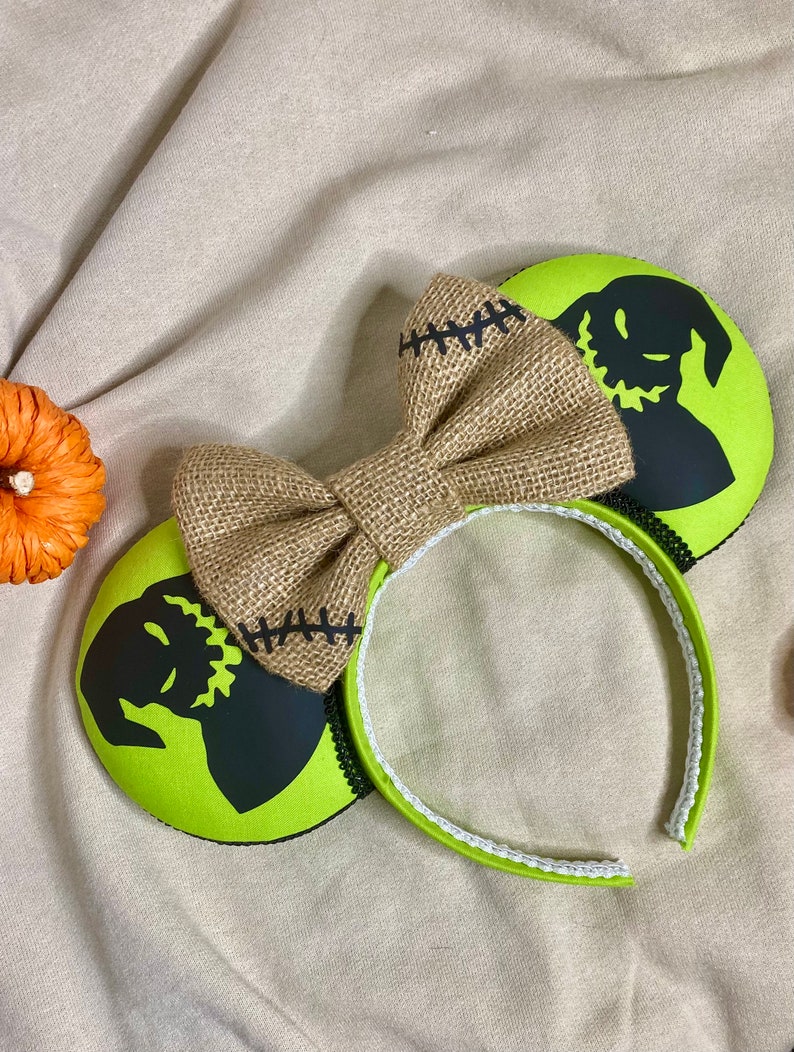 May include: Green and black mouse ears with a burlap bow. The ears feature a silhouette of the character Jack Skellington from the movie The Nightmare Before Christmas.