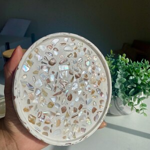 May include: A round, white decorative tray with a mosaic design. The mosaic is made of iridescent, shell-like pieces in shades of white, beige, and brown. The tray is held by a hand and is near a small potted plant.