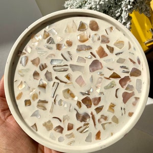 May include: A circular, cream-coloured tray featuring a mosaic pattern. The mosaic is made up of iridescent shell fragments in cream, tan, and brown. The tray has a raised edge, and the diameter is approximately 15 cm.