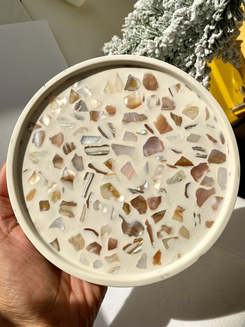 May include: A circular, cream-coloured tray featuring a mosaic pattern. The mosaic is made up of iridescent shell fragments in cream, tan, and brown. The tray has a raised edge, and the diameter is approximately 15 cm.