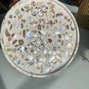 May include: A round, white decorative tray with a terrazzo design. The tray is embedded with iridescent shell fragments in shades of white, brown, and iridescent colours. The tray has a slightly raised edge.