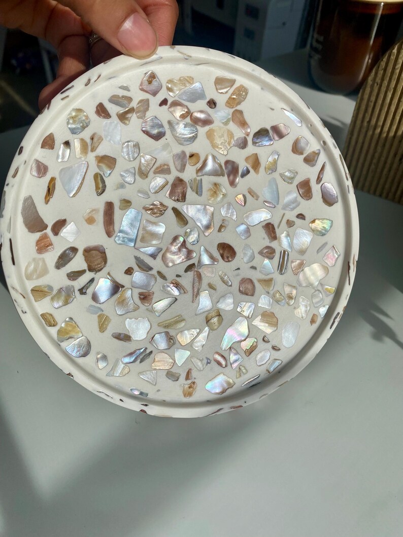 May include: A round, white decorative tray with a terrazzo design. The tray is embedded with iridescent shell fragments in shades of white, brown, and iridescent colours. The tray has a slightly raised edge.