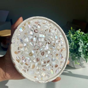 May include: A circular, white decorative tray featuring a mosaic design. The mosaic is composed of iridescent, shell-like fragments in white, brown, and gold tones. The tray is held in a hand, with a small plant in a marble pot visible in the background.