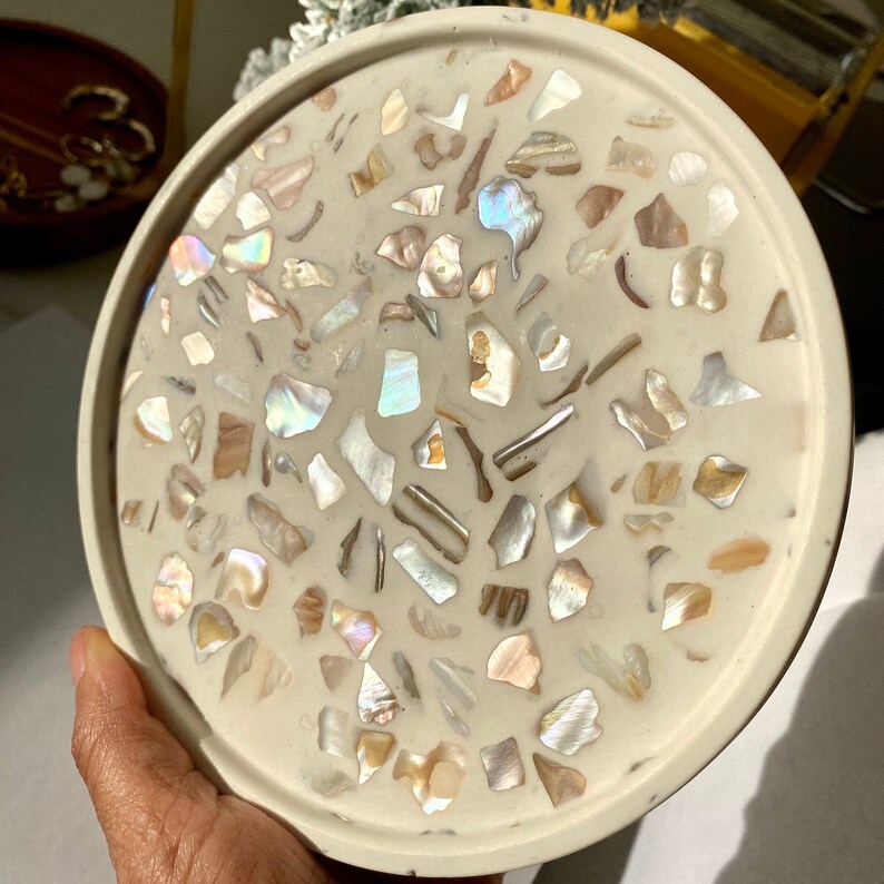 May include: A circular, cream-coloured decorative tray featuring a terrazzo pattern. The tray is inlaid with iridescent shell pieces in white, beige, and brown hues. The tray's edge is a solid cream colour.