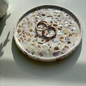 May include: A round, off-white decorative tray with a mosaic of iridescent shell pieces. Two gold hoop earrings rest on the tray. The tray is approximately 15 cm in diameter, ideal for jewelry or small items.