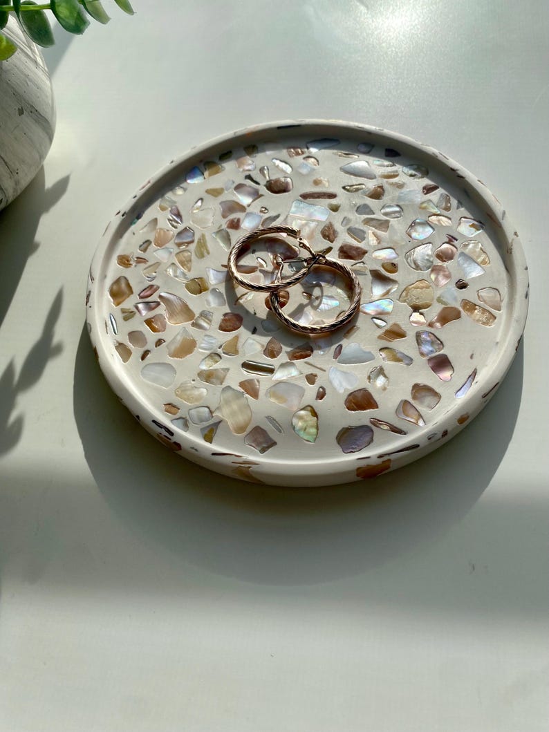 May include: A round, off-white decorative tray with a mosaic of iridescent shell pieces. Two gold hoop earrings rest on the tray. The tray is approximately 15 cm in diameter, ideal for jewelry or small items.