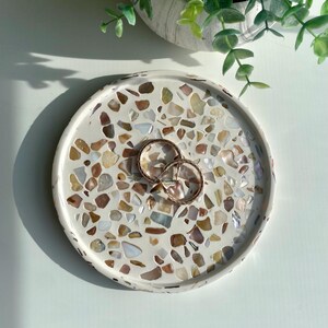 May include: A circular, white decorative tray featuring a mosaic pattern of brown, beige, and cream-coloured stones. Two gold hoop earrings are placed on the tray. A small, white pot with green plants is in the background.