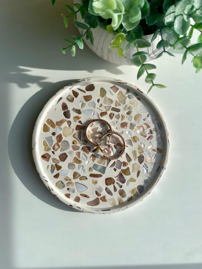 May include: A circular, white decorative tray featuring a mosaic pattern of brown, beige, and cream-coloured stones. Two gold hoop earrings are placed on the tray. A small, white pot with green plants is in the background.