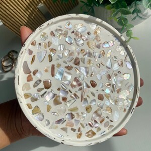 May include: A round, white decorative tray with a mosaic design. The mosaic is made of iridescent shell pieces in shades of white, brown, and gold. The tray is held in a hand.