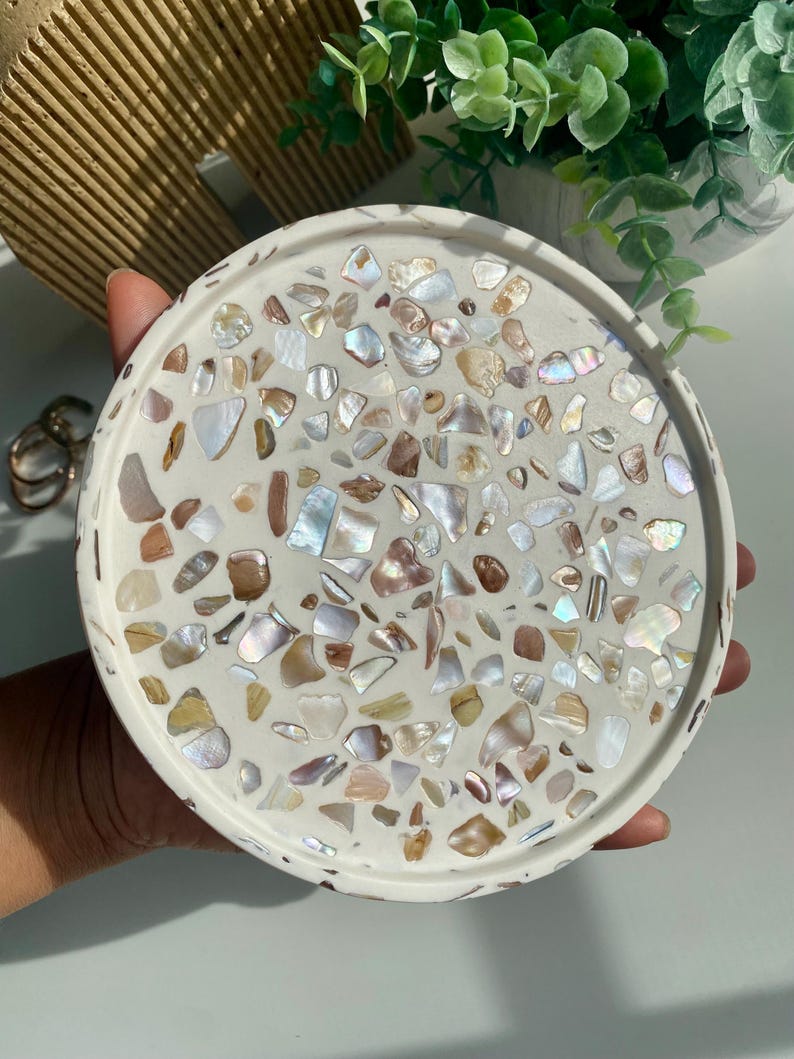 May include: A round, white decorative tray with a mosaic design. The mosaic is made of iridescent shell pieces in shades of white, brown, and gold. The tray is held in a hand.
