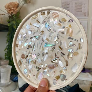 May include: A circular resin tray in off-white, embedded with iridescent shell pieces. The tray has a raised edge and a glossy finish. The shell fragments are of various sizes and shapes, creating a unique decorative surface. A handmade item.