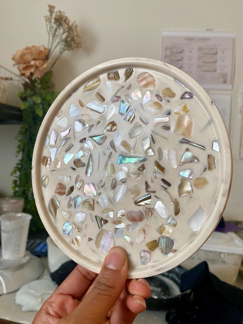 May include: A circular resin tray in off-white, embedded with iridescent shell pieces. The tray has a raised edge and a glossy finish. The shell fragments are of various sizes and shapes, creating a unique decorative surface. A handmade item.