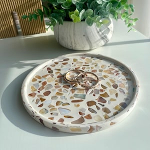 May include: A circular, white terrazzo tray featuring a mosaic of colourful stone chips. Two gold hoop earrings are placed on the tray. The tray is a decorative item, suitable for holding jewellery or small items.