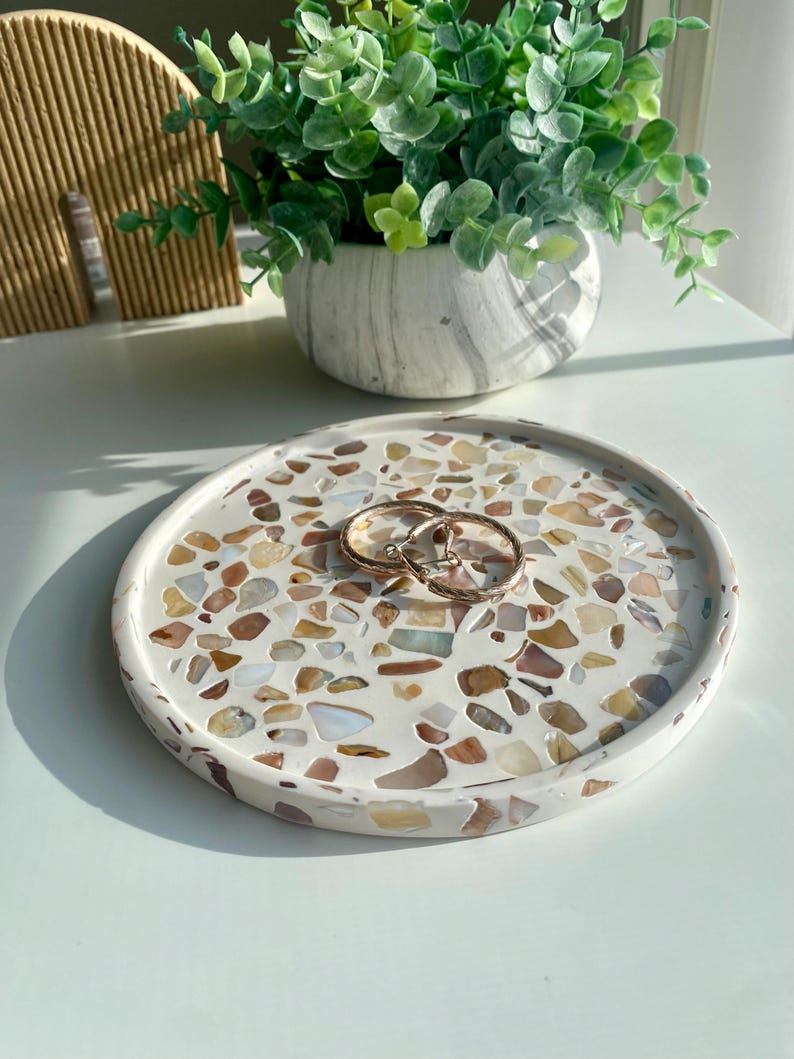 May include: A circular, white terrazzo tray featuring a mosaic of colourful stone chips. Two gold hoop earrings are placed on the tray. The tray is a decorative item, suitable for holding jewellery or small items.