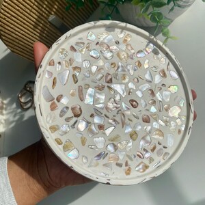 May include: A circular, off-white decorative tray with a mosaic design. The mosaic is made of iridescent shell pieces in white, beige, and brown. The tray is held by a person's hand.
