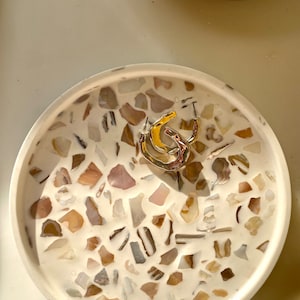 May include: A circular, white tray featuring a mosaic pattern of beige, brown, and grey stones. A gold and silver ring is in the centre. A wooden bowl with gold jewellery is in the background.