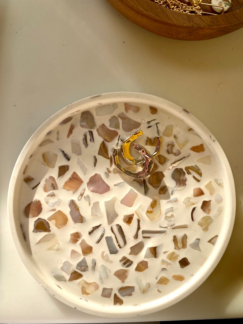 May include: A circular, white tray featuring a mosaic pattern of beige, brown, and grey stones. A gold and silver ring is in the centre. A wooden bowl with gold jewellery is in the background.